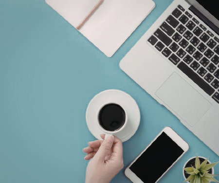 A hand holding a coffee cup on a blue desk with a laptop, notebook, smartphone, and plant.
