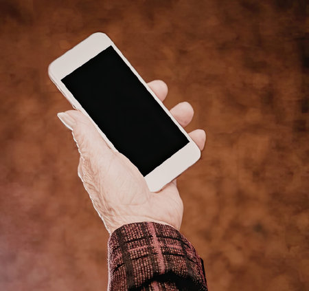 A hand holding a white smartphone with a black screen, wearing a red and black plaid shirt, against...