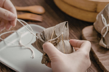 A person is holding a traditional food item wrapped in a leaf, tied with string, on a wooden table...