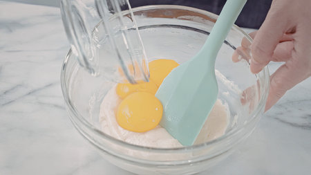 A hand holding a spatula and whisk mixing egg yolks in a glass bowl on a marble countertop.
