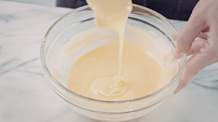 A hand pouring a yellow liquid into a clear glass bowl on a white marble countertop.