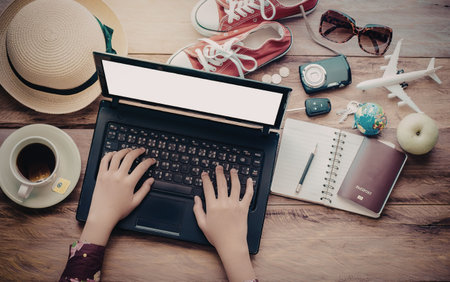 A woman's hands on a laptop keyboard with travel related items on a wooden table