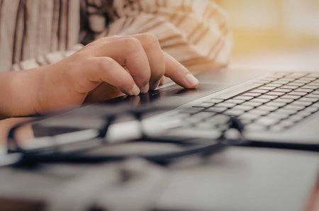 Close-up of a person's hand typing on a laptop keyboard with glasses on the desk