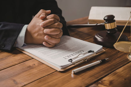A person wearing a suit sits at a wooden desk with their hands clasped together in front of them....