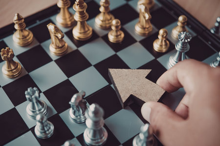 A hand holding a wooden arrow shaped chess piece on a chessboard with gold and silver pieces.
