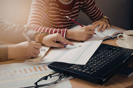 Two individuals collaborating on documents with a laptop, glasses, and coffee on the table.