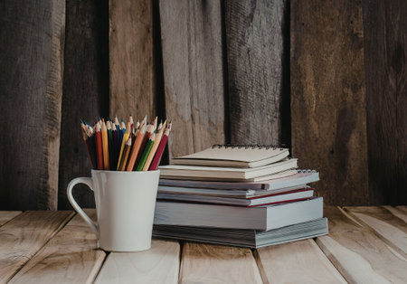 A stack of books and a mug filled with colored pencils on a wooden table