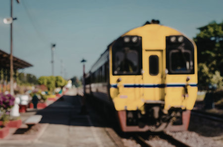 A yellow train is stopped at a railway station with a platform and trees in the background.