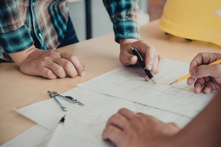 Two people's hands working on a blueprint with a pencil and pen, with a yellow hard hat in the...