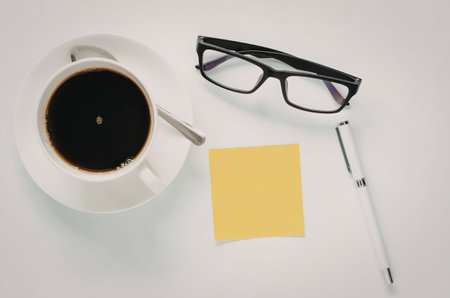 A cup of coffee, glasses, a pen, and a sticky note on a white deskの写真素材