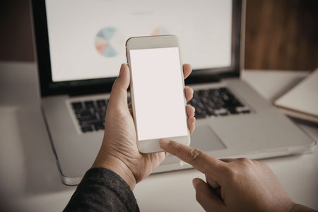 A person holding a smartphone with a white screen in front of a laptop on a desk.