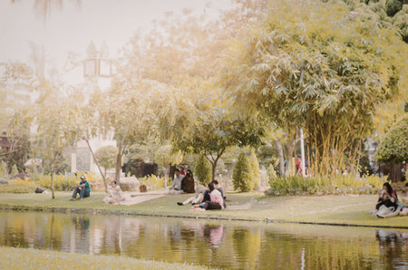 a blurry image of people sitting by a pond in a park with trees and grassの写真素材