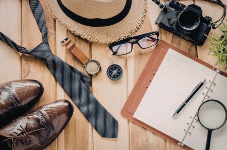 A collection of travel planning essentials including a hat, tie, shoes, camera, glasses, watch, notebook, pen, and magnifying glass arranged on a wooden table.の写真素材