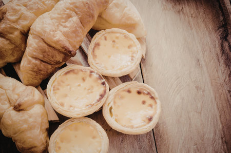An assortment of golden-brown croissants and egg tarts on a rustic wooden surface