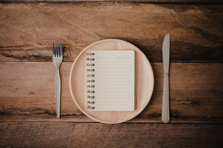 A wooden plate with a spiral-bound notepad, fork, and knife on a rustic wooden table.の写真素材