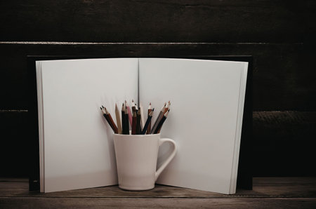 A white mug filled with colored pencils sitting on a wooden desk in front of two pieces of paper.