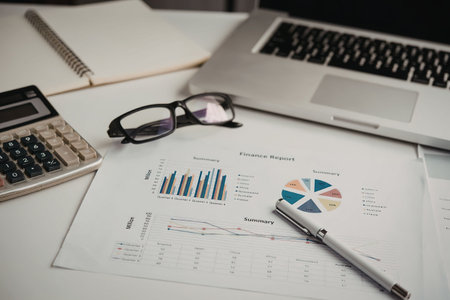 A workspace with a laptop, calculator, glasses, and papers with graphs and charts, indicating a...
