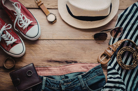 A flat lay of casual summer clothing and accessories including a hat, sneakers, jeans, belt, wallet, sunglasses, headphones, and a striped shirt.の写真素材