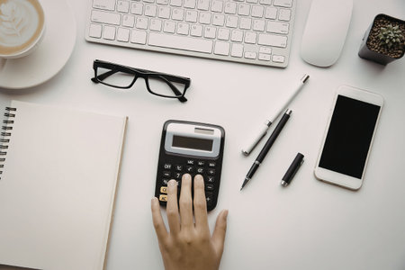 A hand on a calculator with office supplies on a white desk.