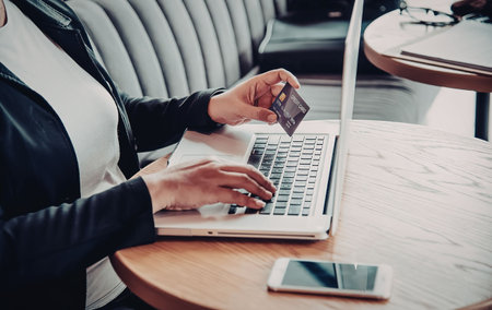 A woman sitting at a table using a laptop and holding a credit card, likely shopping online.の写真素材