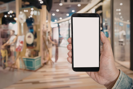 A hand holding a smartphone with a blank white screen in a shopping mall background.の写真素材