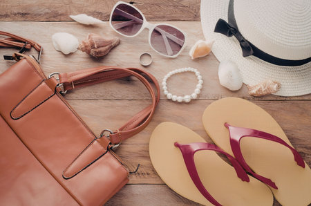 A collection of summer vacation accessories including a leather bag, sunglasses, jewelry, flip flops, and a sun hat, arranged on a wooden surface.の写真素材