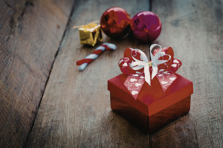 A red gift box with a white ribbon sits on a wooden surface surrounded by Christmas ornaments and a...