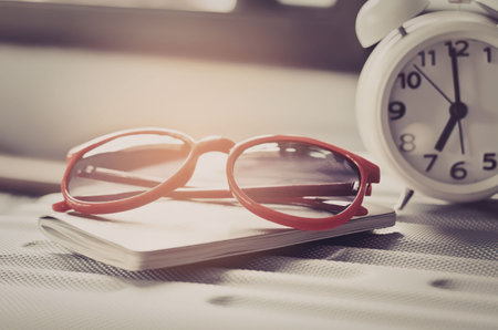 A pair of red glasses on a book next to a white alarm clock
