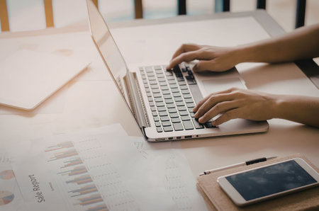A person typing on a laptop with documents and a phone on the desk