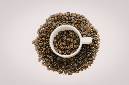 A white cup filled with coffee beans and surrounded by more coffee beans on a plain white...