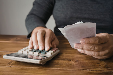 A person sitting at a wooden table, using a calculator and holding a piece of paper.