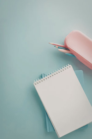 A spiral notebook with a blank white page sits on top of a blue notebook, accompanied by a pink...