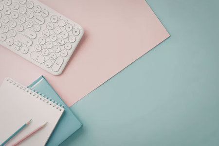 A white keyboard, spiral notebook, colored pencils and pastel colored paper on a blue and pink desk