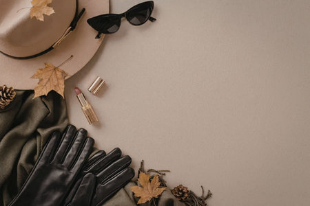 A flat lay of autumn fashion accessories including a hat, sunglasses, gloves, scarf, and makeup on a beige background with fall leaves and pinecones.の写真素材