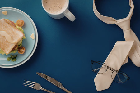 An overhead view of a breakfast setting with a sandwich on a plate, a cup of coffee, a fork, a knife, a pair of glasses, and a necktie on a blue background.の写真素材