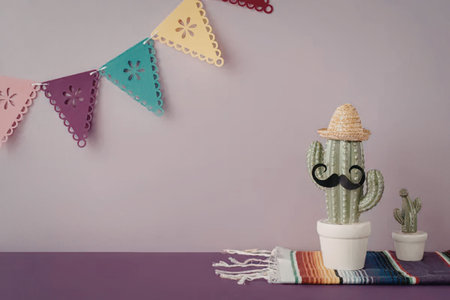 A cactus wearing sunglasses and a hat sits on a colorful blanket next to another small cactus, with a purple wall and a banner of colorful triangular flags in the background.の写真素材