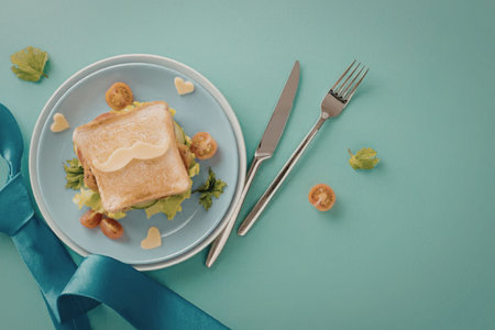 A sandwich on a plate with a knife and fork, surrounded by cherry tomatoes and herbs on a light blue background.の写真素材