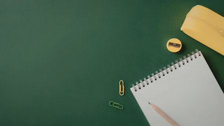 A spiral notebook, pencil, eraser, paperclip and pencil sharpener on a green background