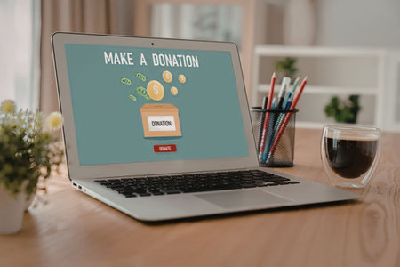 Laptop computer on wooden desk displays make a donation screen with charity box and coins, accompanied by a cup of coffee, promoting online giving and charitable donations.の写真素材