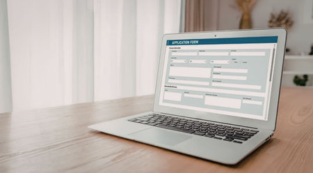 Modern silver laptop on wooden desk near window displays online application form on its screen in a...