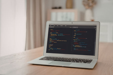 Modern laptop sits on a wooden table displaying coding software in a well lit office environment...