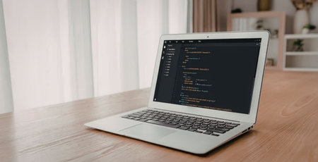 Modern silver laptop on a wooden table displaying lines of code on its dark screen in a bright and...