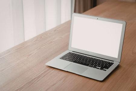 Modern silver laptop with a blank white screen sits on a wooden table in a bright office setting,...