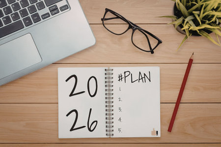 Notebook on a wooden desk displays 2026 and a plan with a laptop, glasses, pencil, and plant...