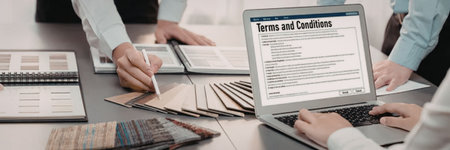 Professionals gather around table reviewing terms and conditions on laptop screen with design...