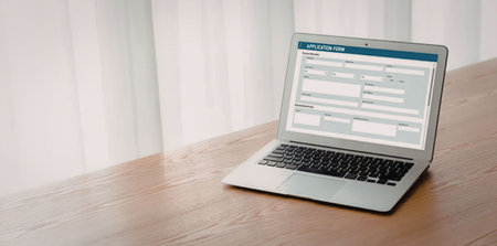 Silver laptop on a wooden desk displaying an online application form with various fields to be...