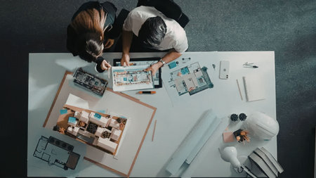 Two architects collaborate on a design project with blueprints and models on a white table, engaging...