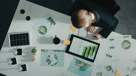 A businessman in a black suit sits at a white table analyzing green energy graphs on a tablet...
