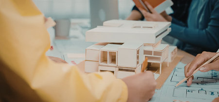 a group of people sitting around a table working on a model of a building, one person wearing yellow is holding the model while another person in blue is looking at blueprintsの写真素材