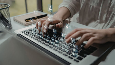 A person typing on a laptop keyboard with a notebook and glass nearby